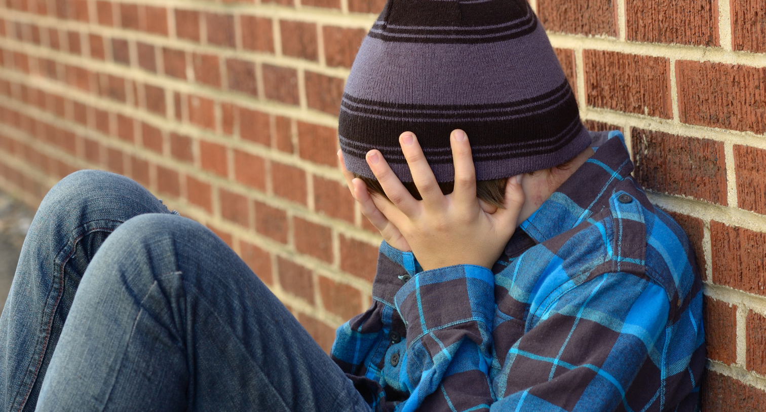 Teenage boy sitting down outside leaning on brick wall with his head in his hands