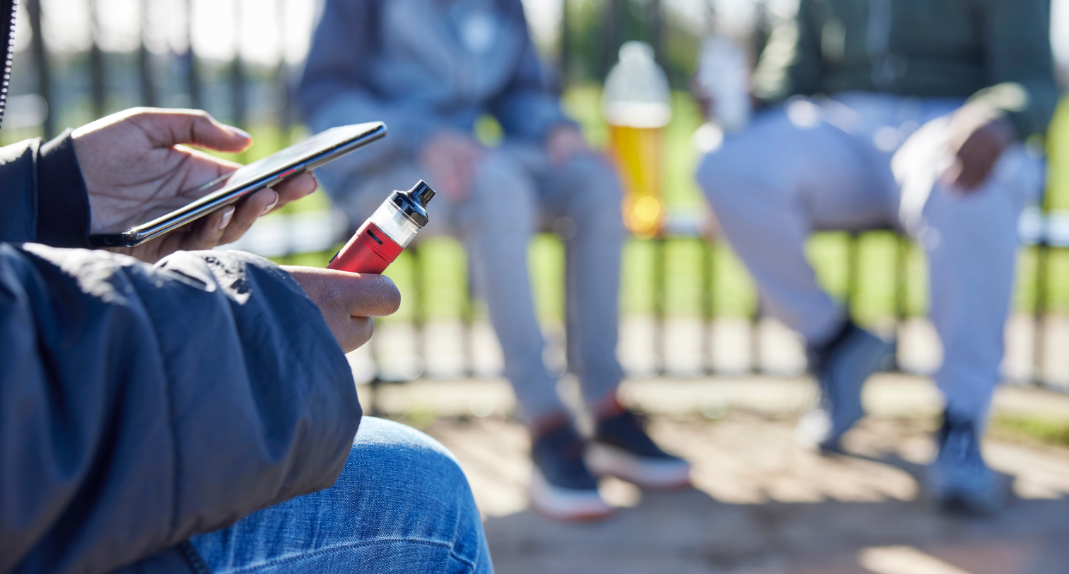 Close Up Of Teenagers With Mobile Phone Vaping and Drinking Alcohol In Park