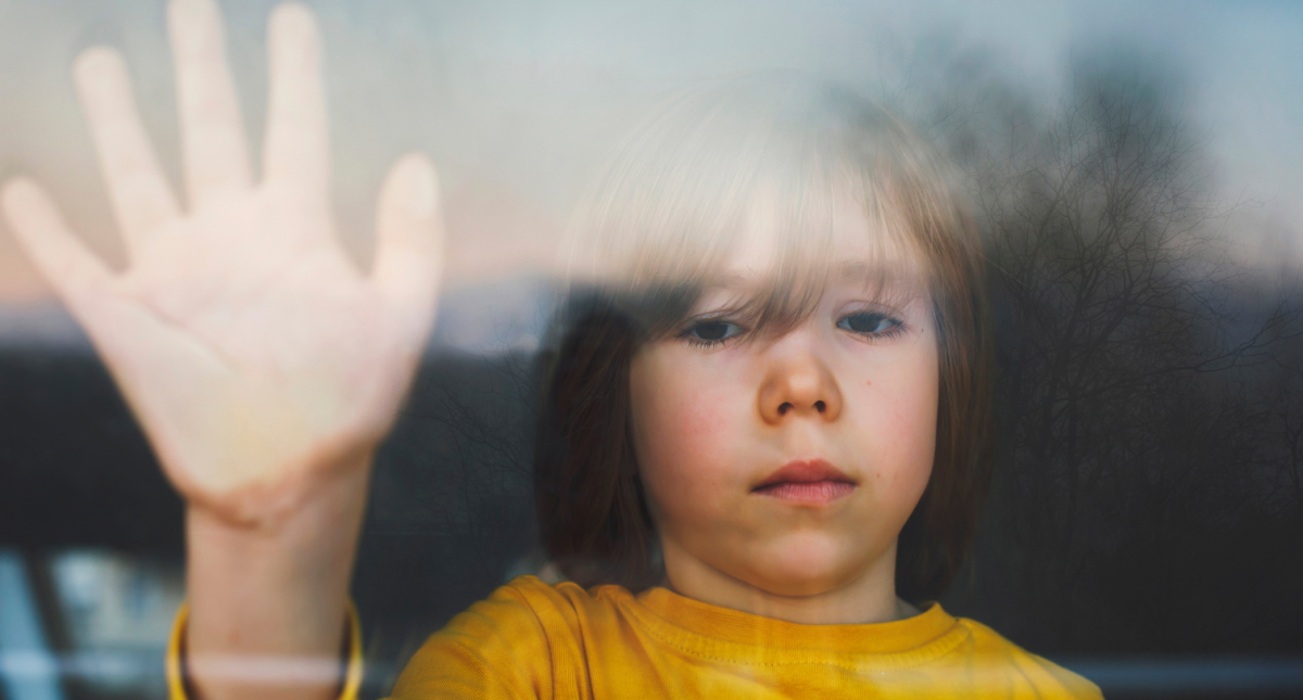 A child looking out a window with a hand on the window looking sad
