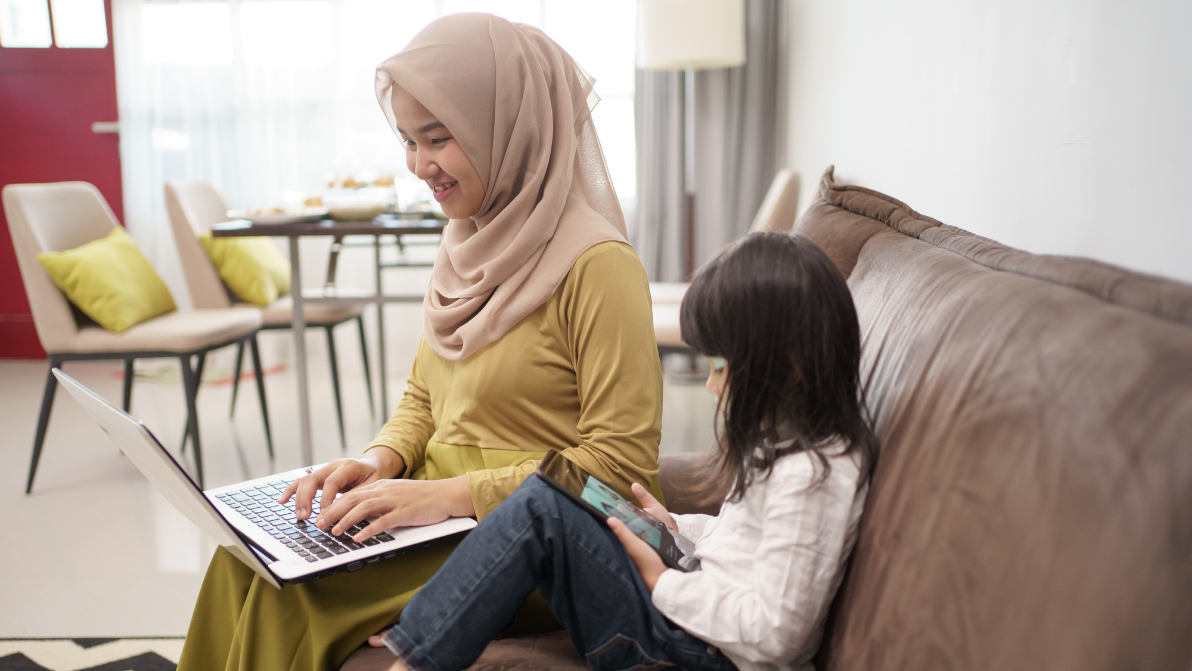 Mother taking online parenting course on a laptop, sitting on sofa with her daughter.